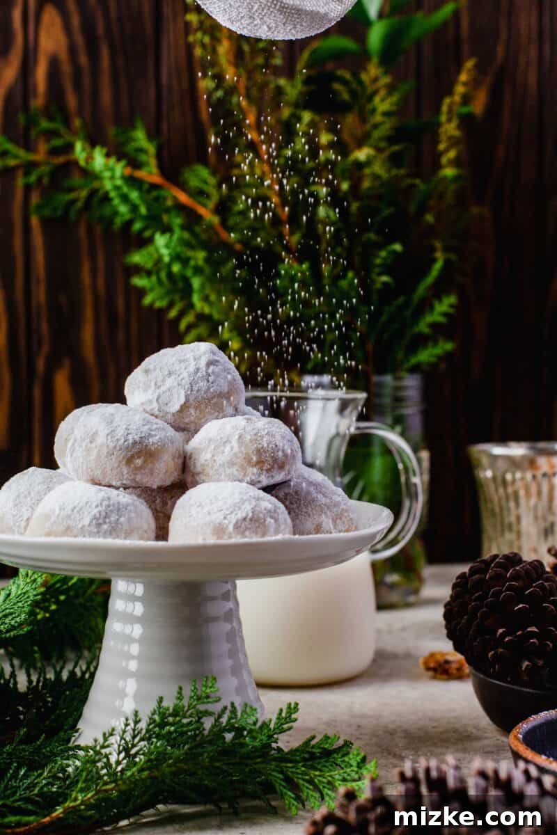 Melt-in-Your-Mouth Snowballs 2 a stack of snowball cookies on a cake stand with powdered sugar being sprinkled above. pinecones in a bowl can be seen next to the cake stand