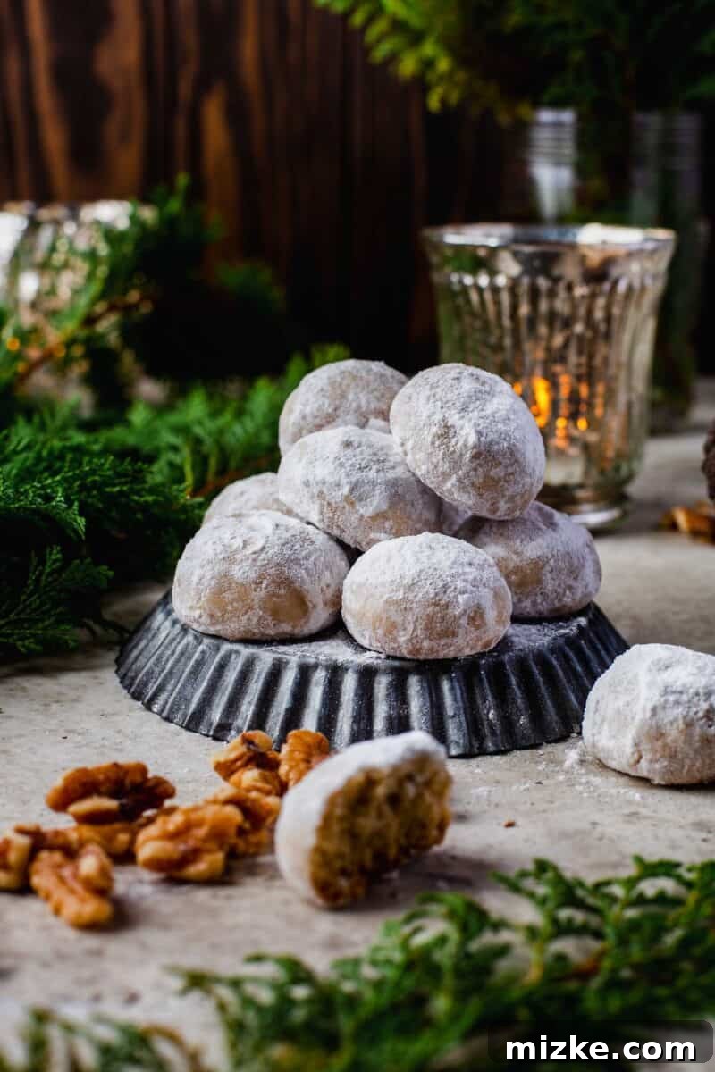 Melt-in-Your-Mouth Snowballs 11 snowball cookies stacked on top of an upside down dark grey tart pan. whole walnuts and evergreen pine leaves can be seen in the scenery surrounding the snowball cookies. a dim candle is lit in the background as well