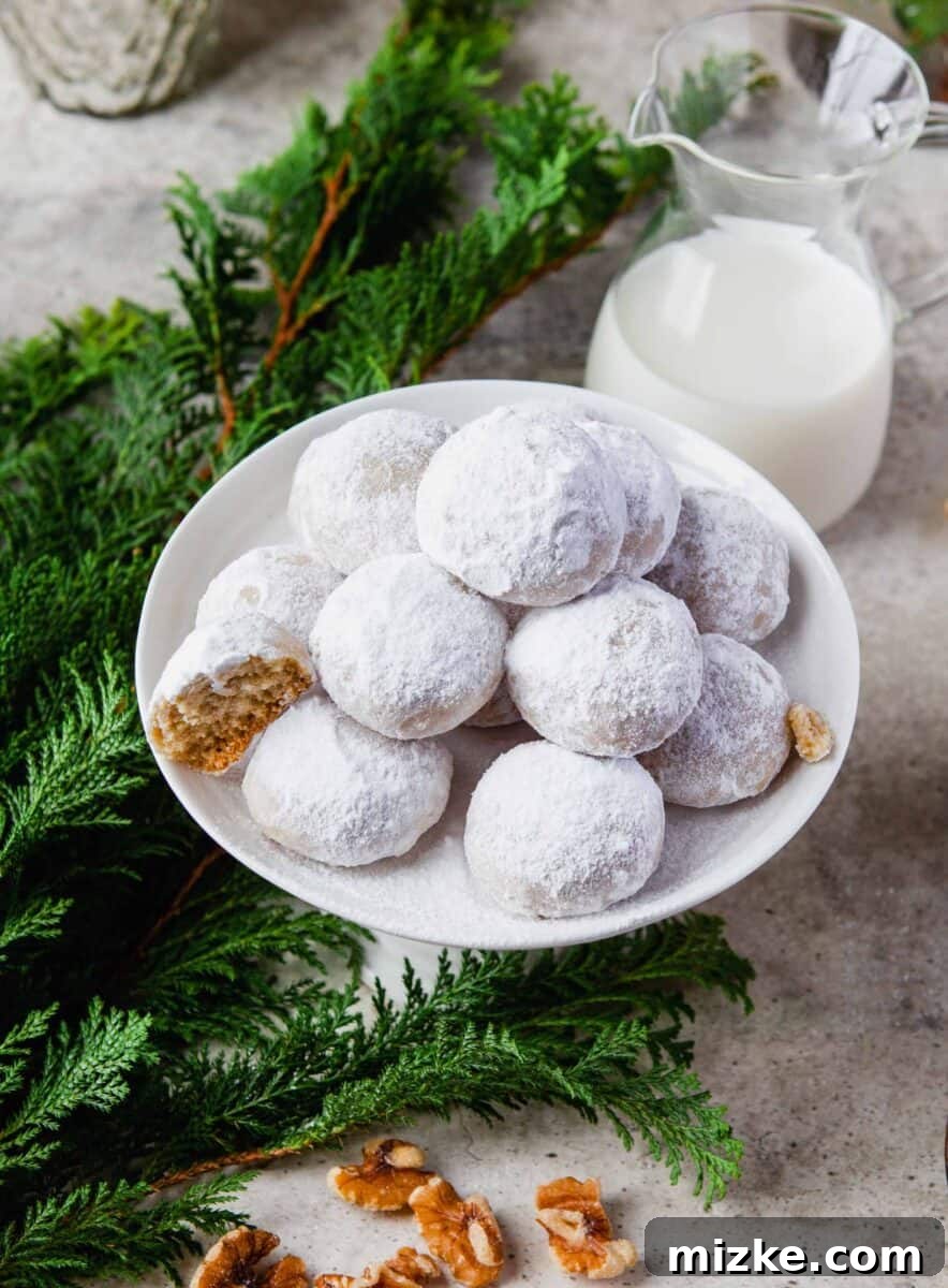 Melt-in-Your-Mouth Snowballs 12 a pile of snowball cookies on a white plate with evergreen tree leaves next to it and a carafe of milk