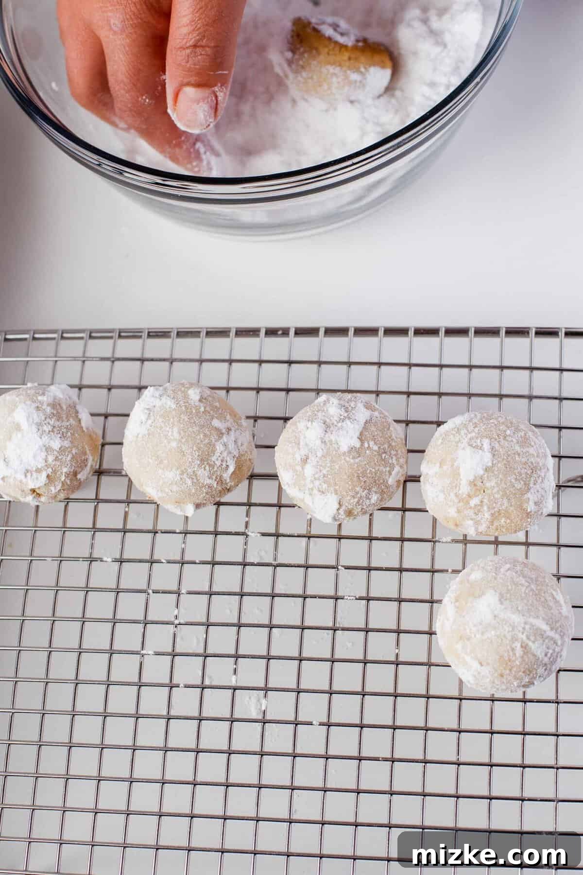 Melt-in-Your-Mouth Snowballs 10 dusted snowball cookies on a metal baking rack with a hand tossing a cookie in powdered sugar in the background
