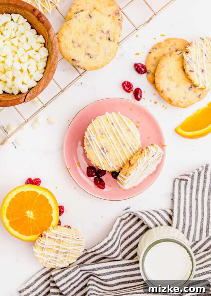 Golden Orange Cranberry Shortbread Delights 13 Overhead shot of a cranberry shortbread cookie on a pink plate next to an orange, a bowl of white chocolate chips, and more cookies. A glass of milk and a linen striped towel are also visible.