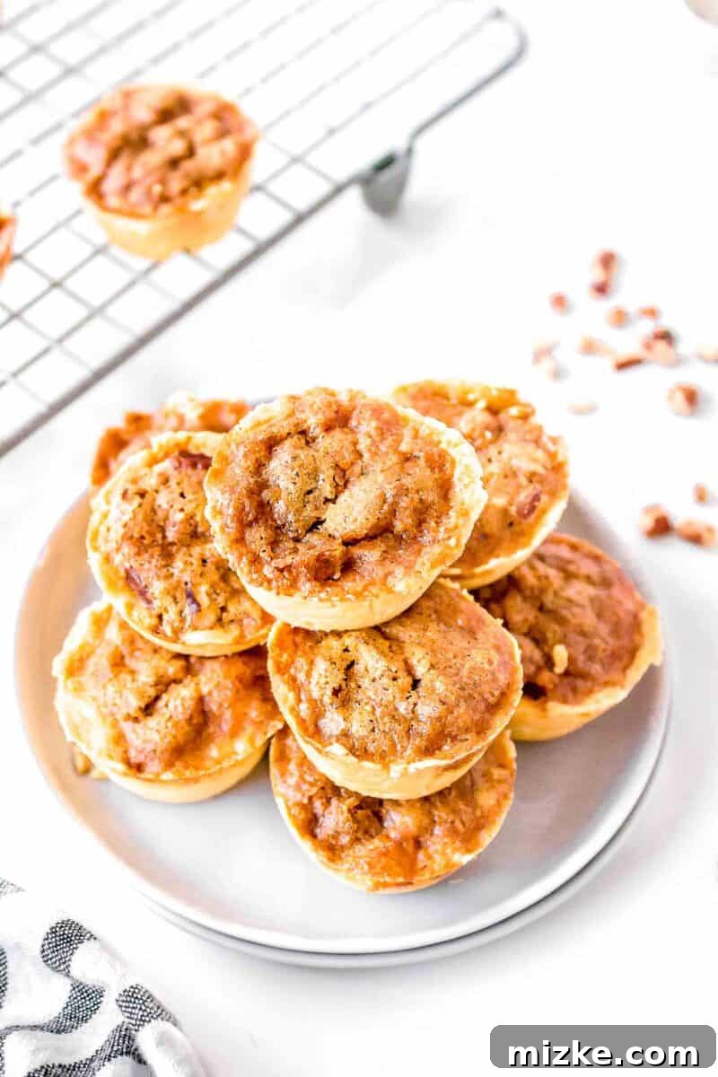 Effortless Pecan Tassie Bites 2 Pecan tassies stacked beautifully on two white plates next to a wire baking rack, showcasing their golden crusts and inviting pecan filling.
