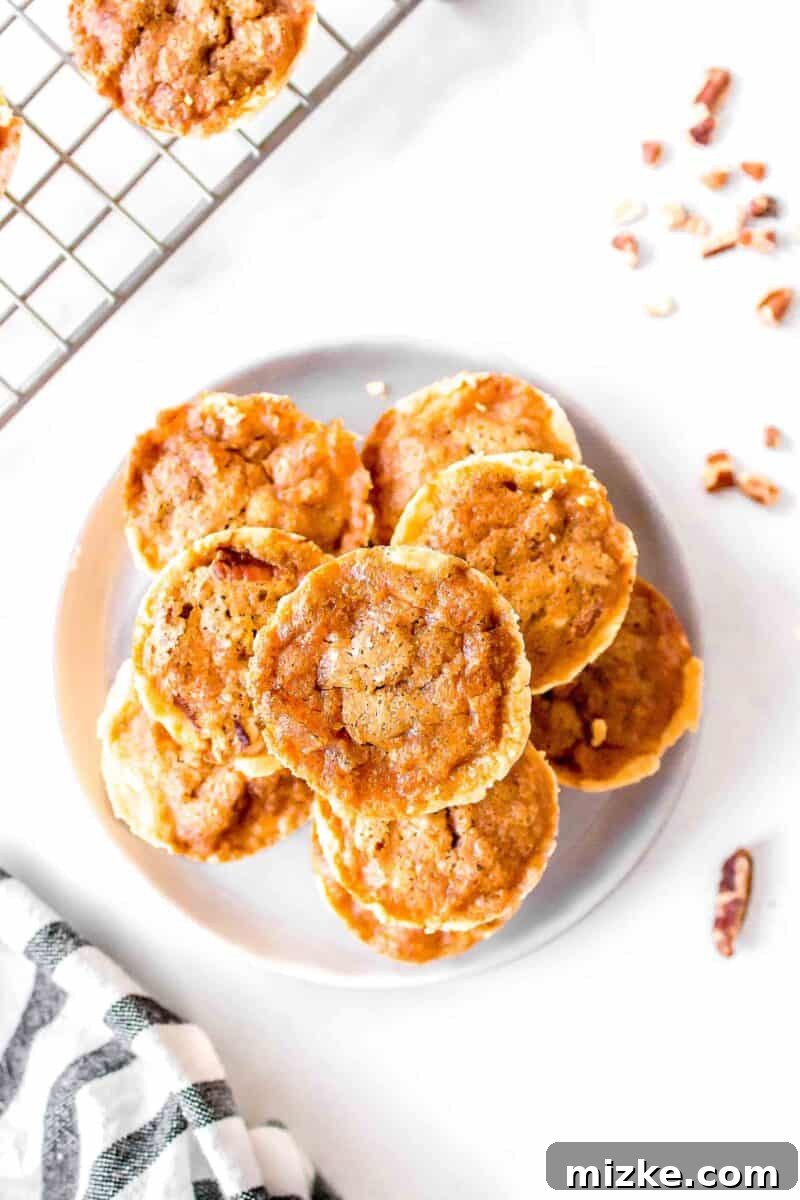 Effortless Pecan Tassie Bites 4 Nine golden-brown pecan tassies perfectly stacked on a small white plate, accompanied by a scattering of crushed pecans and a metal baking rack.