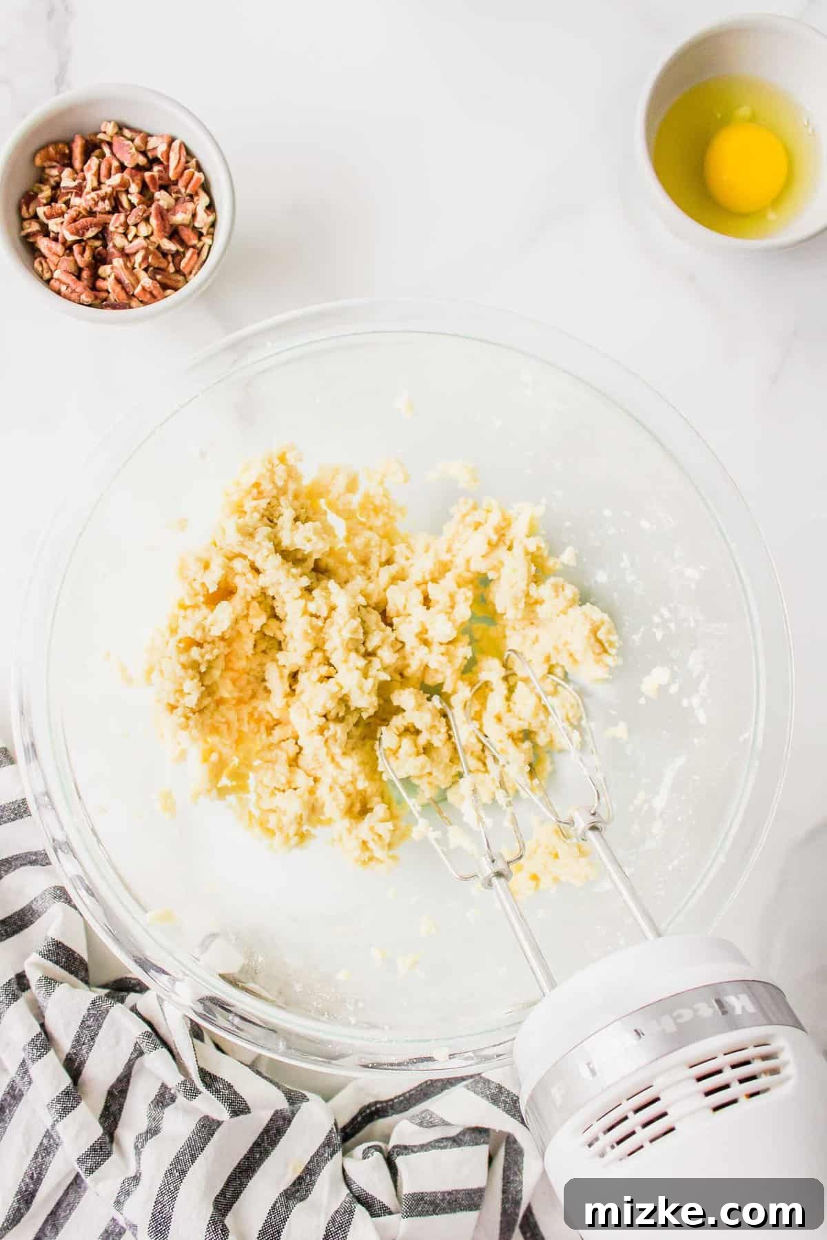 Effortless Pecan Tassie Bites 6 Creaming softened cream cheese, butter, and all-purpose flour together in a clear mixing bowl using a hand mixer, beside a striped linen towel.