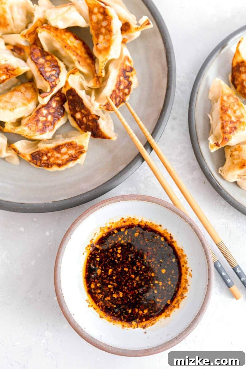 Your New Favorite Dumpling Sauce 5 An inviting overhead view of a brown-rimmed white ceramic bowl filled with our homemade dumpling sauce, flanked by two rustic grey plates piled high with golden-brown, crispy pan-fried dumplings. Elegant wooden chopsticks are gracefully positioned on the plates, ready for dipping.