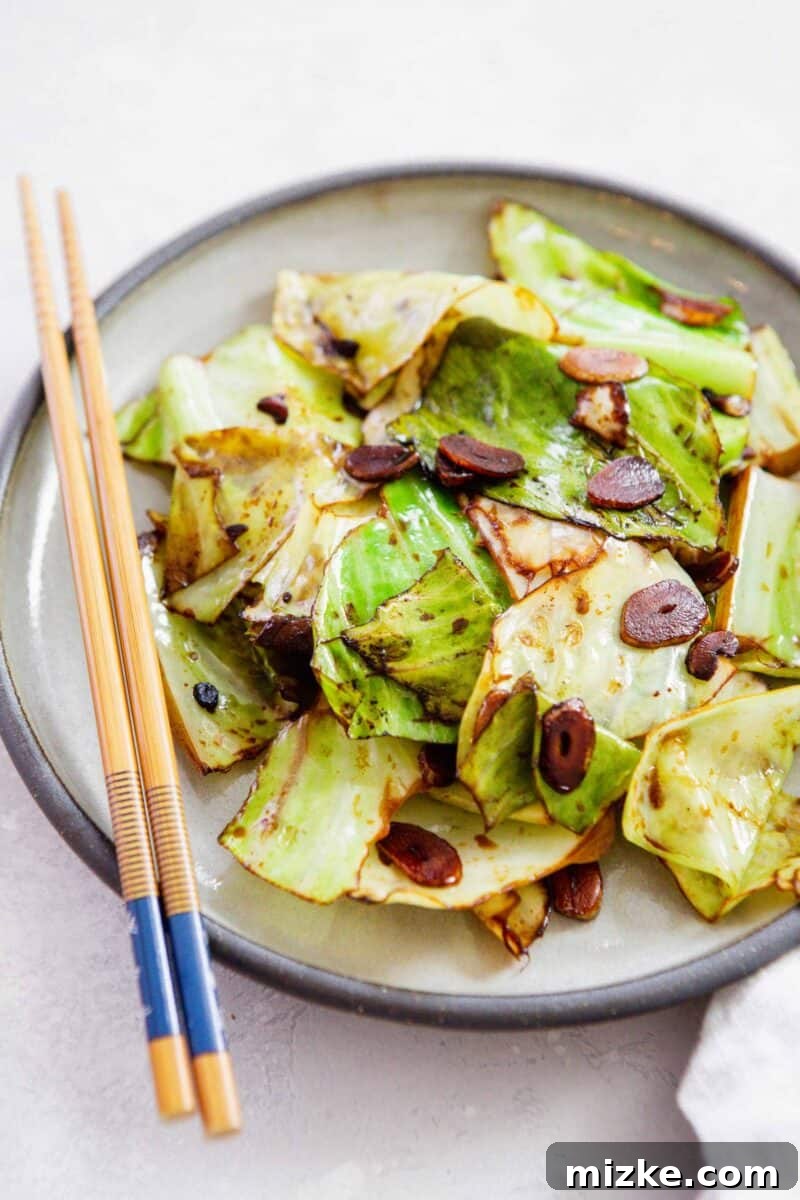 Close-up of stir-fried cabbage tossed in soy sauce with prominent garlic slices, presented on a white plate with wooden chopsticks resting on top.