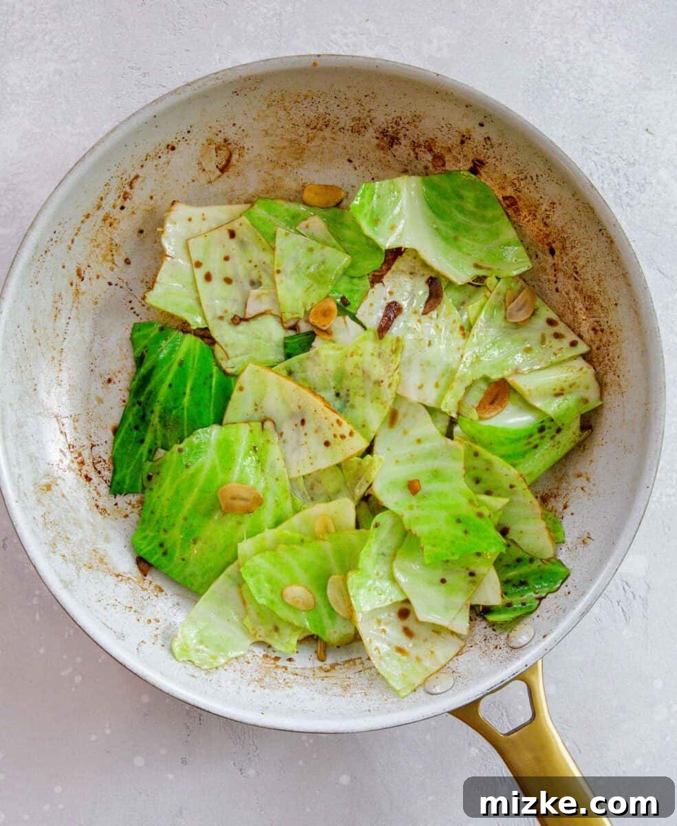 Green cabbage being stir-fried in a hot skillet with garlic slices and a splash of soy sauce, showing the leaves beginning to soften and char slightly.
