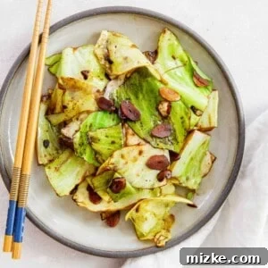 Stir-fried cabbage on a ceramic grey plate with soy sauce coating the cabbage leaves and garlic slices. Wooden chopsticks are visible on the plate and next to a linen towel.