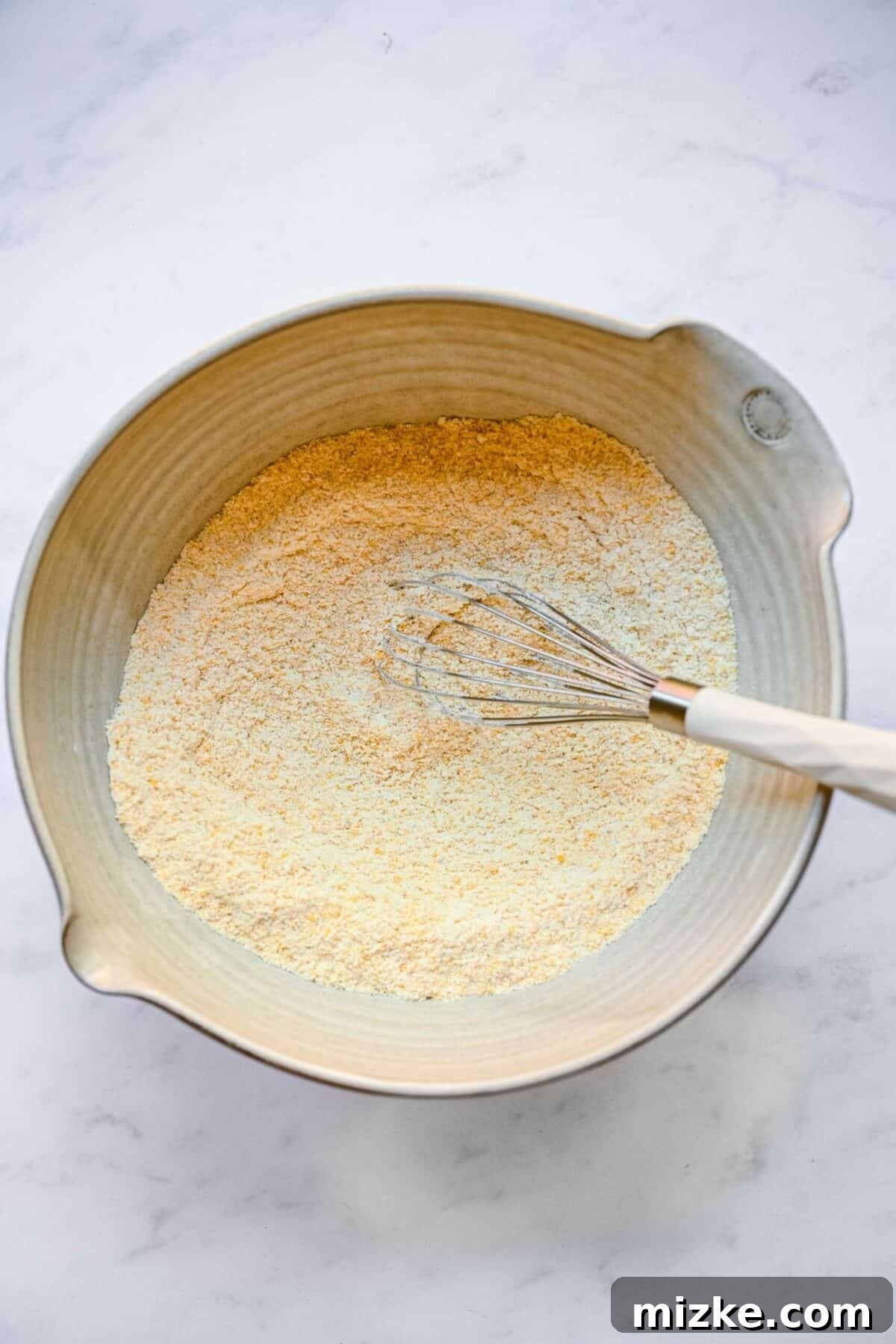 Moist, tender, sweet, and tart, these cranberry maple cornbread muffins are a perfect side for Thanksgiving or Christmas. A mixing bowl containing a well-whisked cornmeal and flour mixture, with a whisk resting inside, signaling readiness for the wet ingredients.