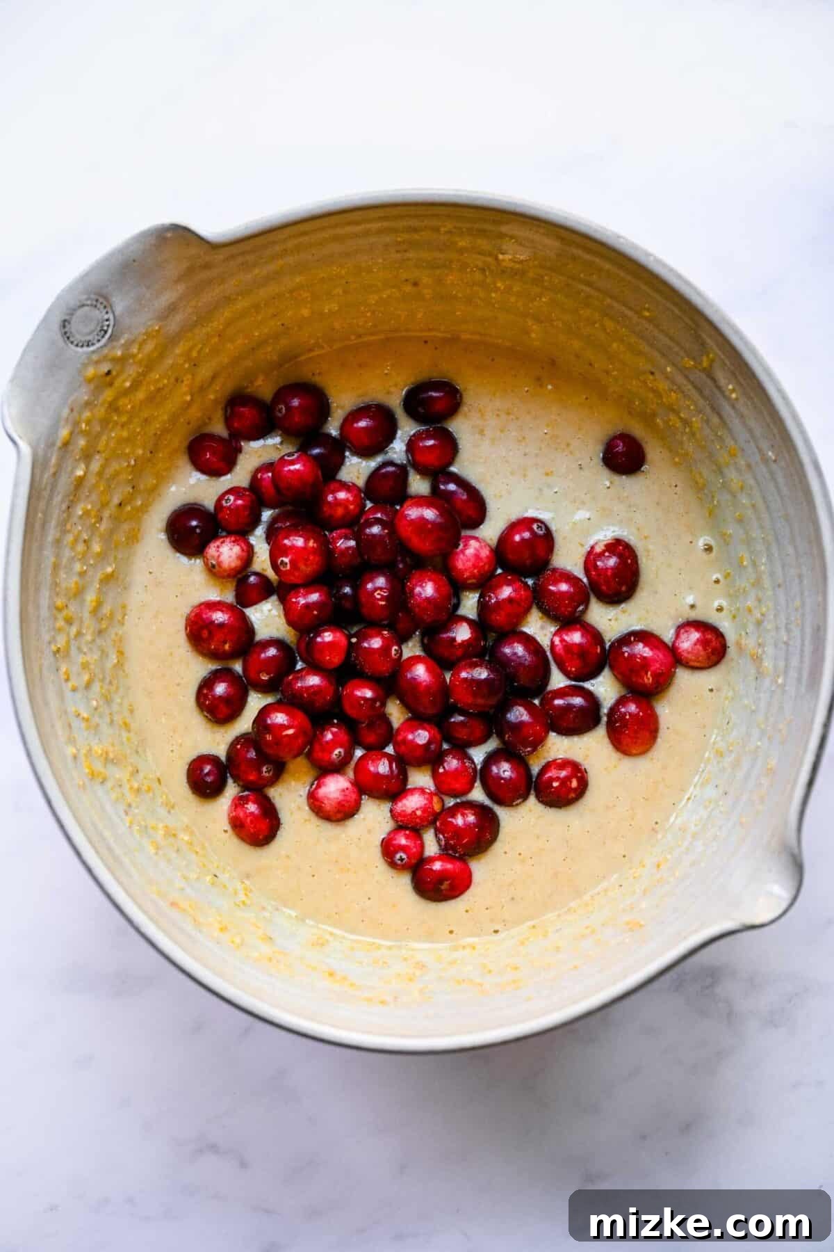 These cranberry maple cornbread muffins are moist, tender, sweet, and tart, making them a superb side dish for Thanksgiving or Christmas. A mixing bowl of golden cornbread muffin batter generously topped with vibrant fresh cranberries, poised for their gentle integration.