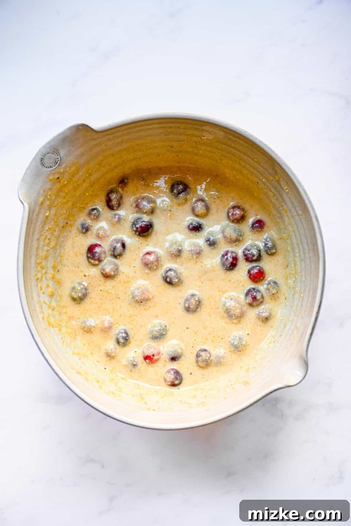 Perfectly moist, tender, sweet, and tart cranberry maple cornbread muffins are an ideal side dish for Thanksgiving or Christmas. The mixing bowl now shows the cornbread muffin batter with fresh cranberries beautifully folded in, creating a marbled effect of color.