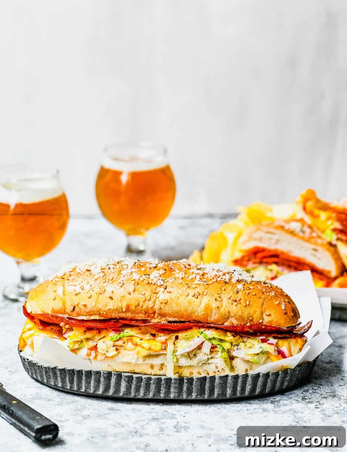 The complete grinder sandwich experience, ready for serving. A large, impressive grinder sandwich on a wooden tray, with two beers and a bag of chips blurred in the background, creating a relaxed meal setting.