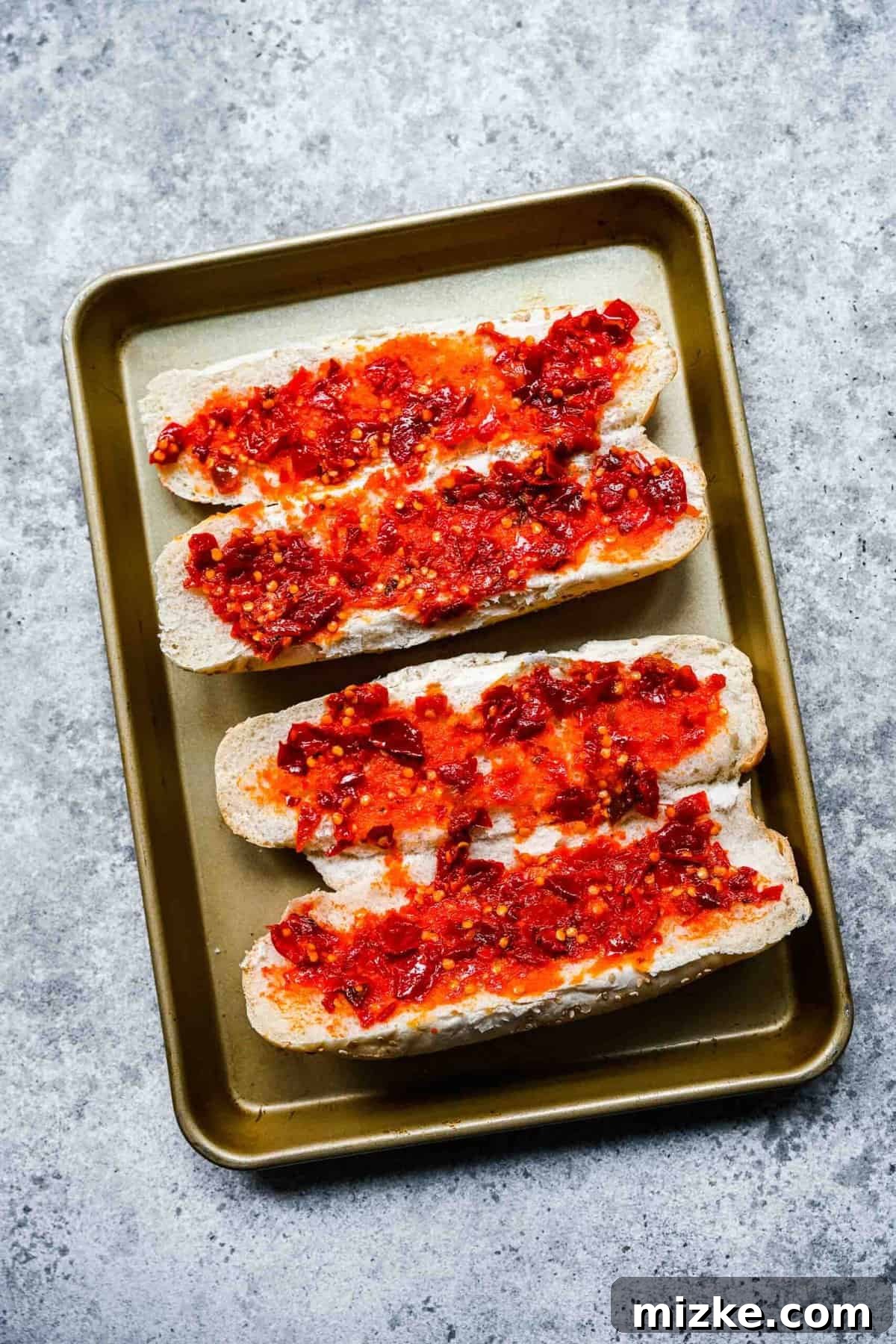 Hoagie rolls prepped with Calabrian chili paste. Two hoagie loaves on a baking sheet, with their interiors coated in spicy Calabrian chili paste, ready for fillings.