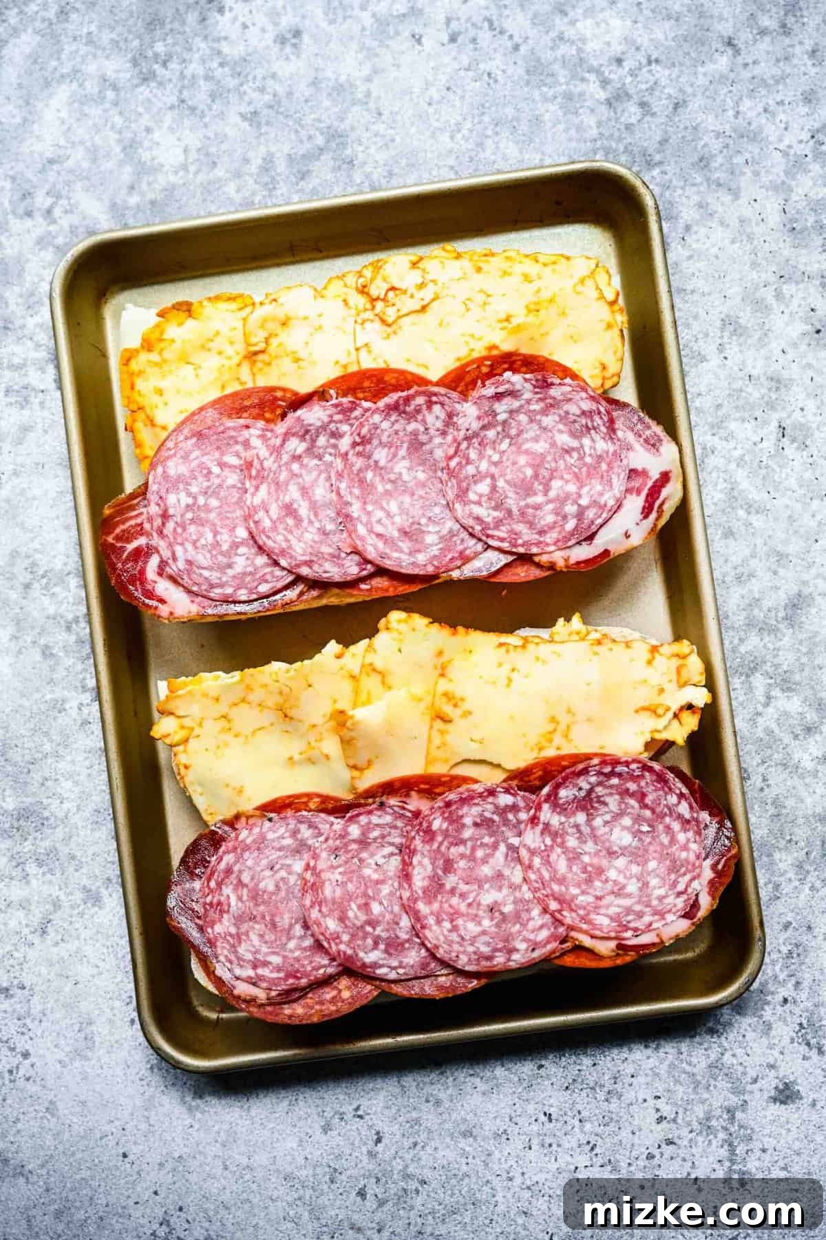 Melted cheese and warmed meats on the hoagie rolls. Two open-faced hoagie rolls on a baking tray after baking, showing melted cheese on one half and warmed cured meats on the other.