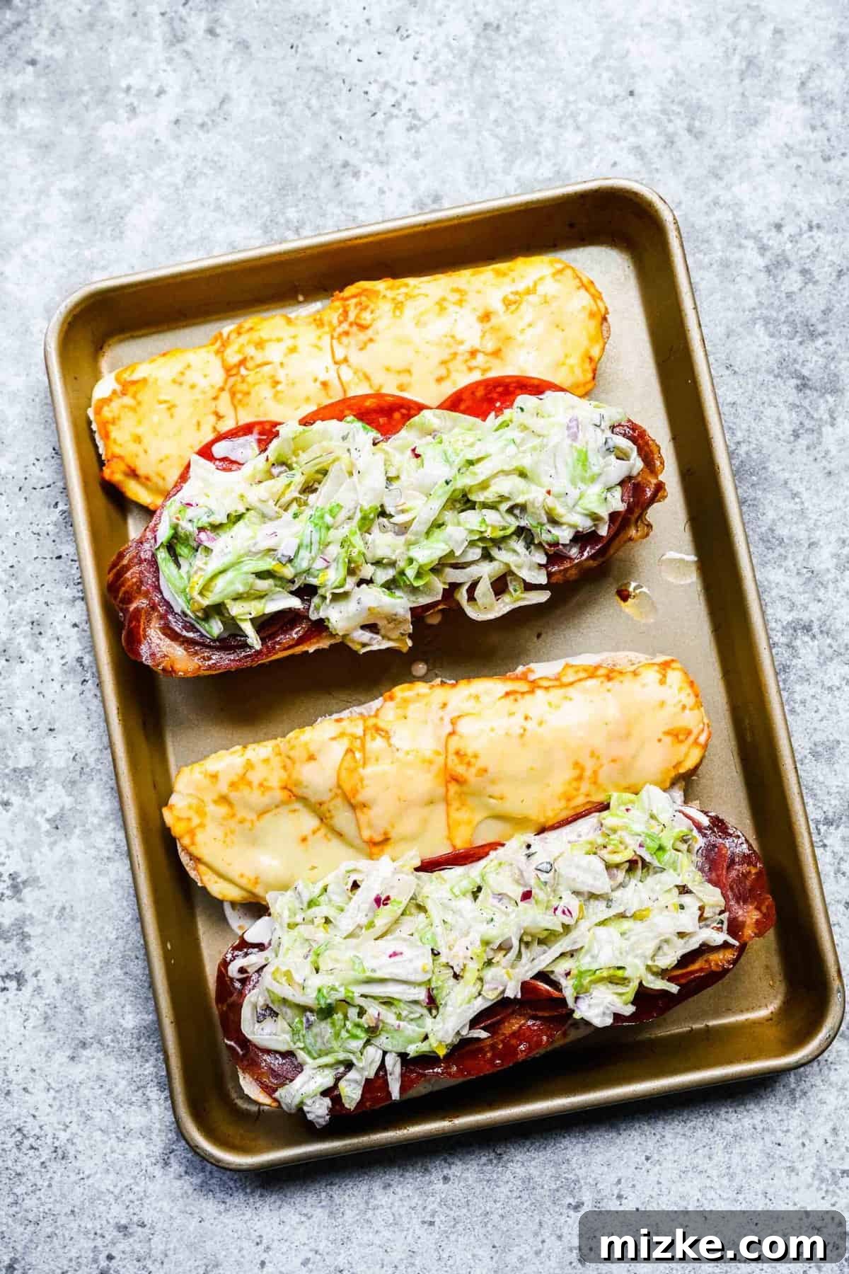 Adding the fresh grinder salad to the warmed meats. Two open hoagie rolls on a baking sheet, with melted cheese on one side and cured meats topped with the fresh grinder salad on the other side.