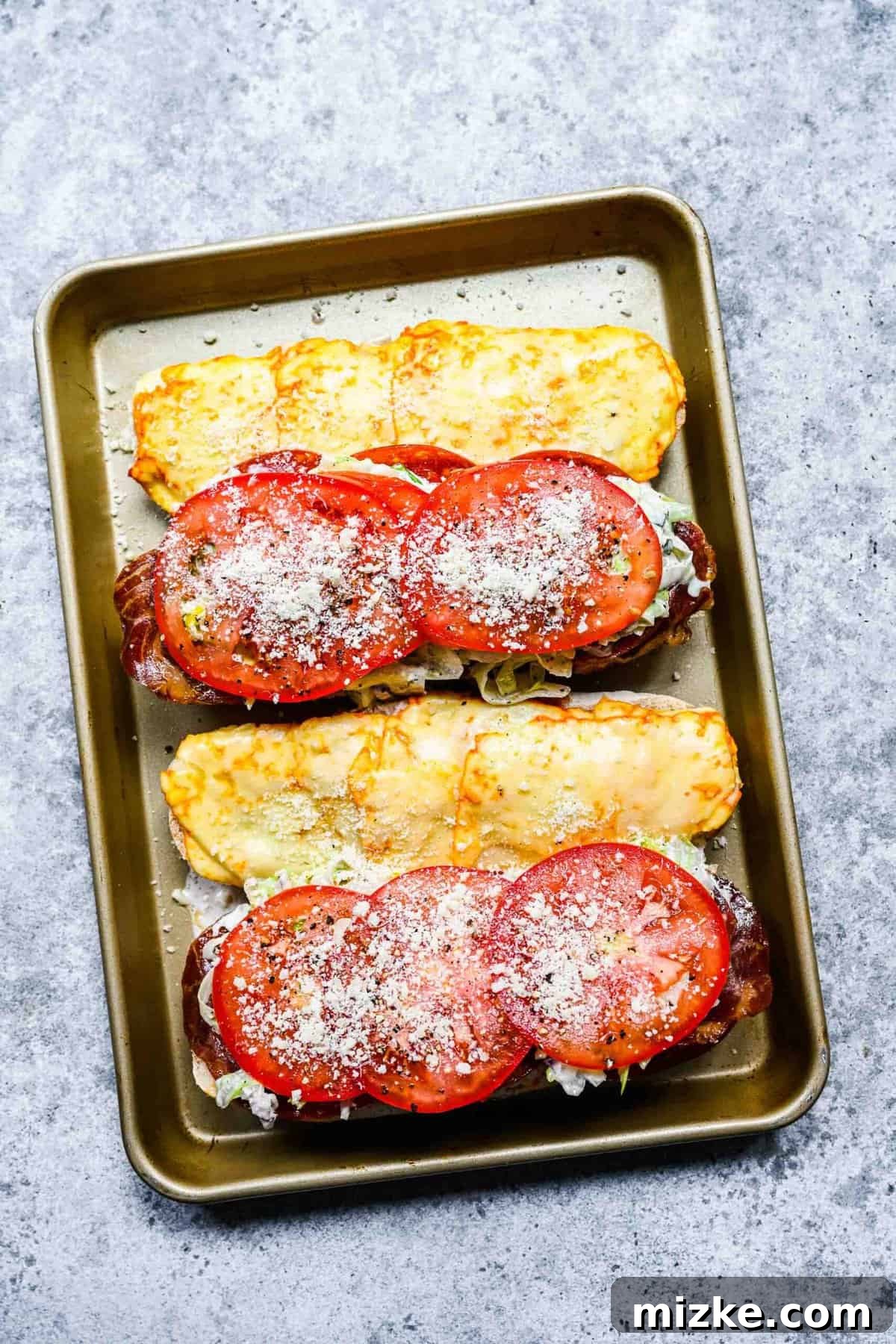 The grinder sandwich, fully loaded with all its fresh ingredients. Two open hoagie loaves on a baking sheet, fully topped with melted cheese, cured meats, salad, tomato slices, salt, pepper, and parmesan.