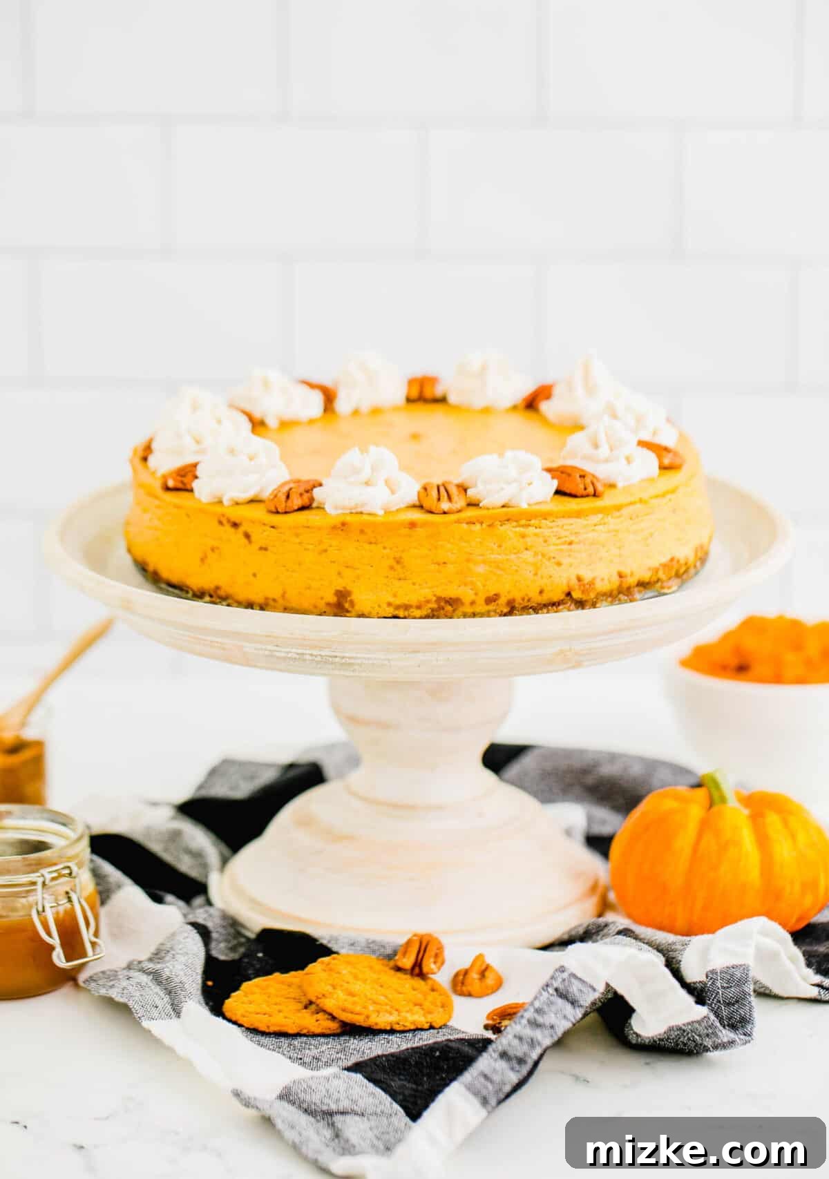 This easy pumpkin cheesecake recipe delivers a rich and creamy homemade cheesecake experience, bursting with authentic pumpkin flavor and infused with cozy fall spices, perfect for any occasion. A beautifully baked pumpkin cheesecake on a cake stand, elegantly topped with candied pecans and swirls of homemade whipped cream, surrounded by decorative mini pumpkins, setting a festive fall scene.