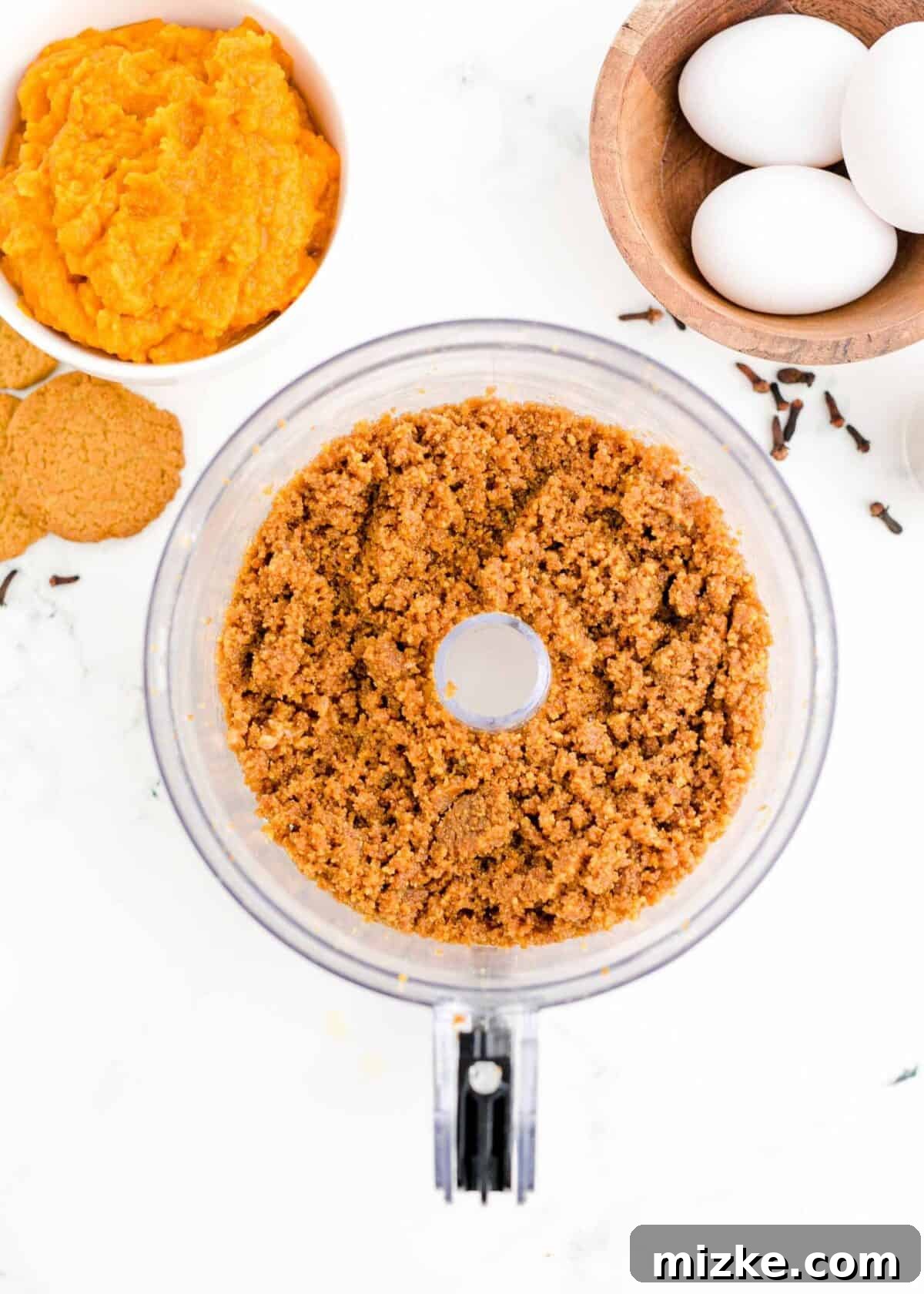 This easy pumpkin cheesecake recipe features a rich and creamy homemade filling, packed with authentic pumpkin flavor and a harmonious blend of cozy fall spices, starting with a perfect gingersnap crust. A food processor bowl filled with finely pulsed gingersnap cookies and melted butter, ready to be pressed into the crust, surrounded by whole cookies, fresh eggs, and a bowl of bright orange pumpkin puree, highlighting the initial steps of cheesecake preparation for a delicious fall dessert.