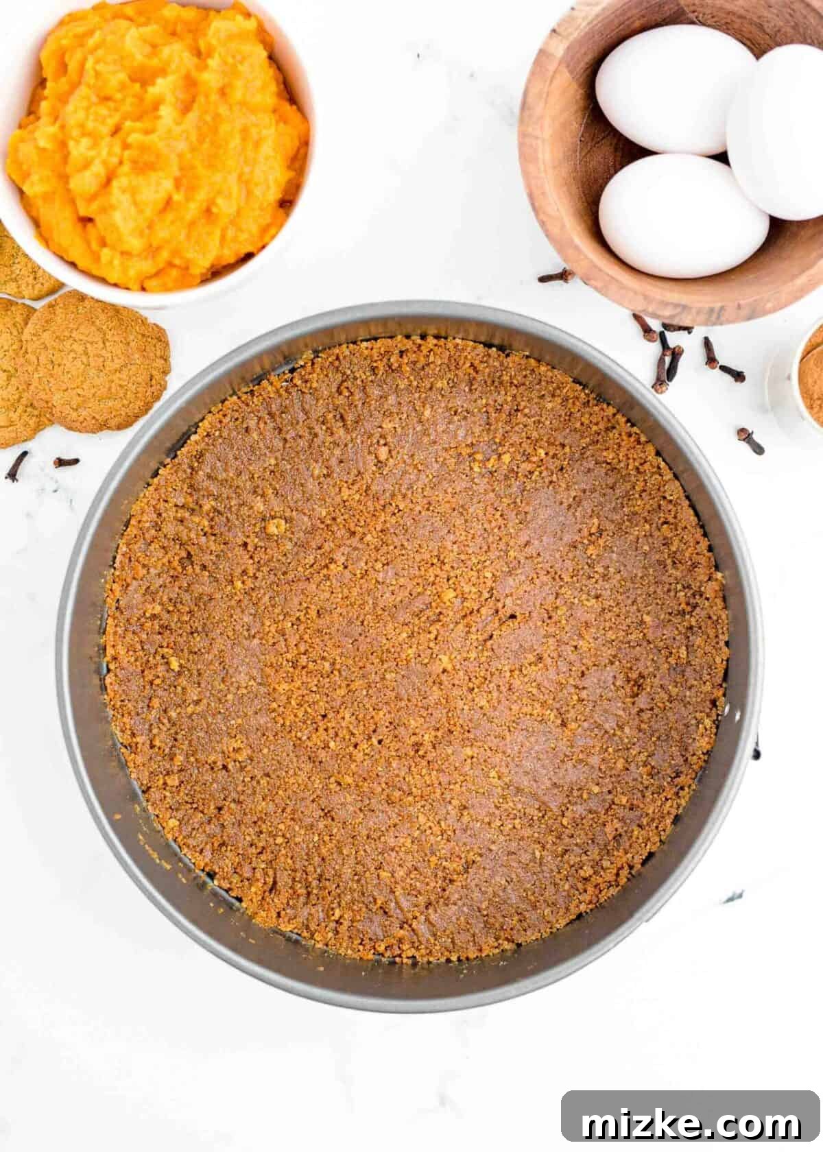 This easy pumpkin cheesecake recipe is beloved for its rich and creamy homemade texture, vibrant with real pumpkin and infused with a captivating mix of fall spices, beginning with a perfectly set crust. The golden-brown gingersnap crust perfectly pressed into the base and slightly up the sides of a greased and parchment-lined springform pan, with additional gingersnaps, eggs, and a bowl of pumpkin puree visible in the background, illustrating the prepared crust before baking.
