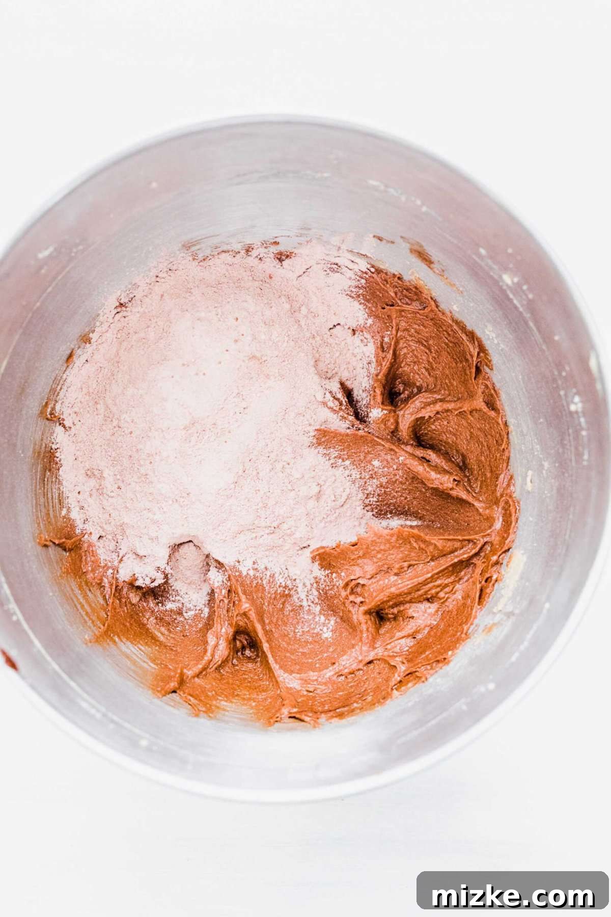 These chocolate peppermint kiss cookies are festive treats for Christmas, easy to make, and topped with a Hershey's candy cane kiss. A mixing bowl holding the combined wet ingredients (creamed butter, sugar, egg, and chocolate), now with a pile of the dry flour and cocoa powder mixture on top, ready for gentle incorporation.