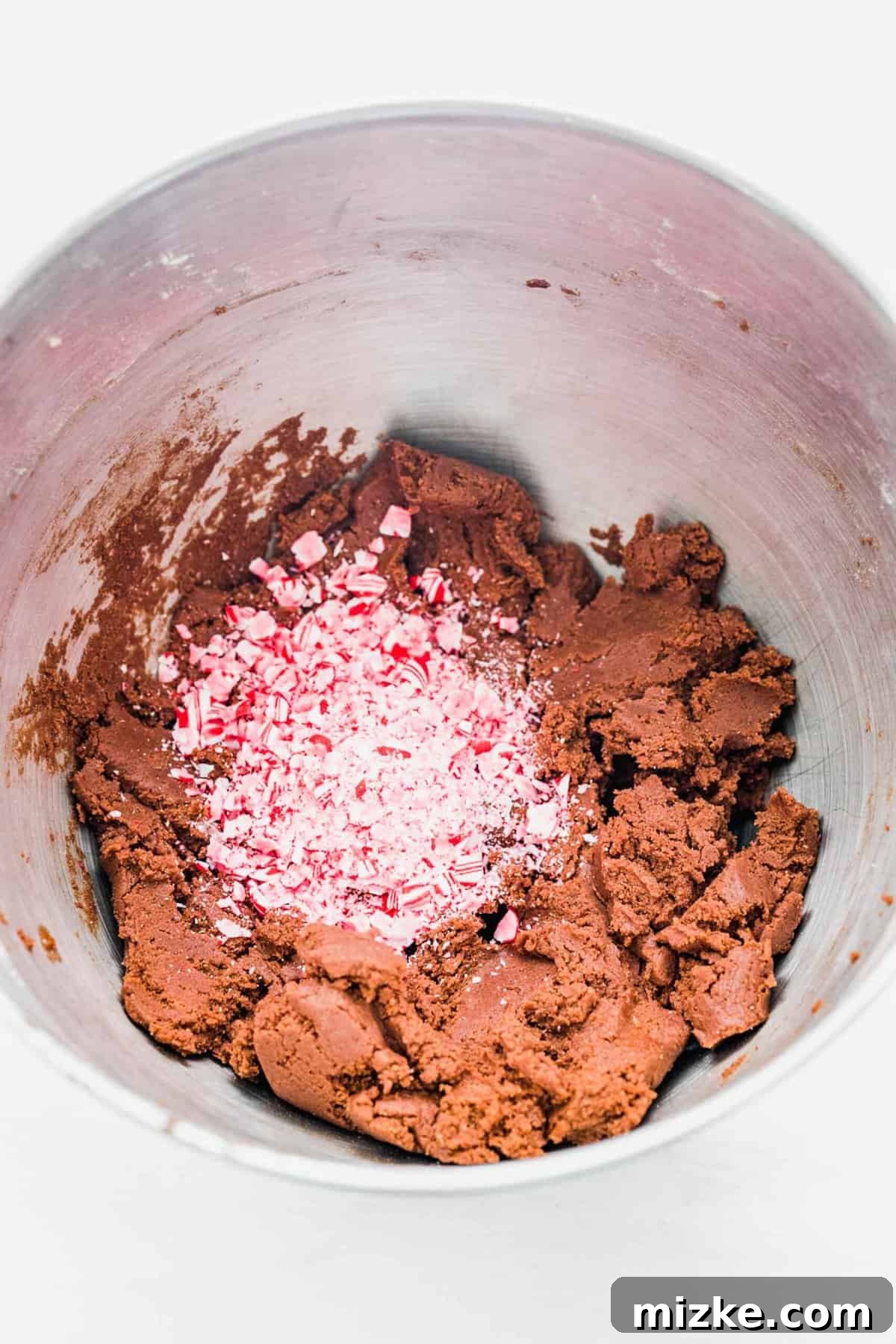These chocolate peppermint kiss cookies are festive treats for Christmas, easy to make, and topped with a Hershey's candy cane kiss. A mixing bowl filled with rich chocolate peppermint cookie dough, with a generous pile of brightly colored crushed candy canes resting on top, ready to be folded in.