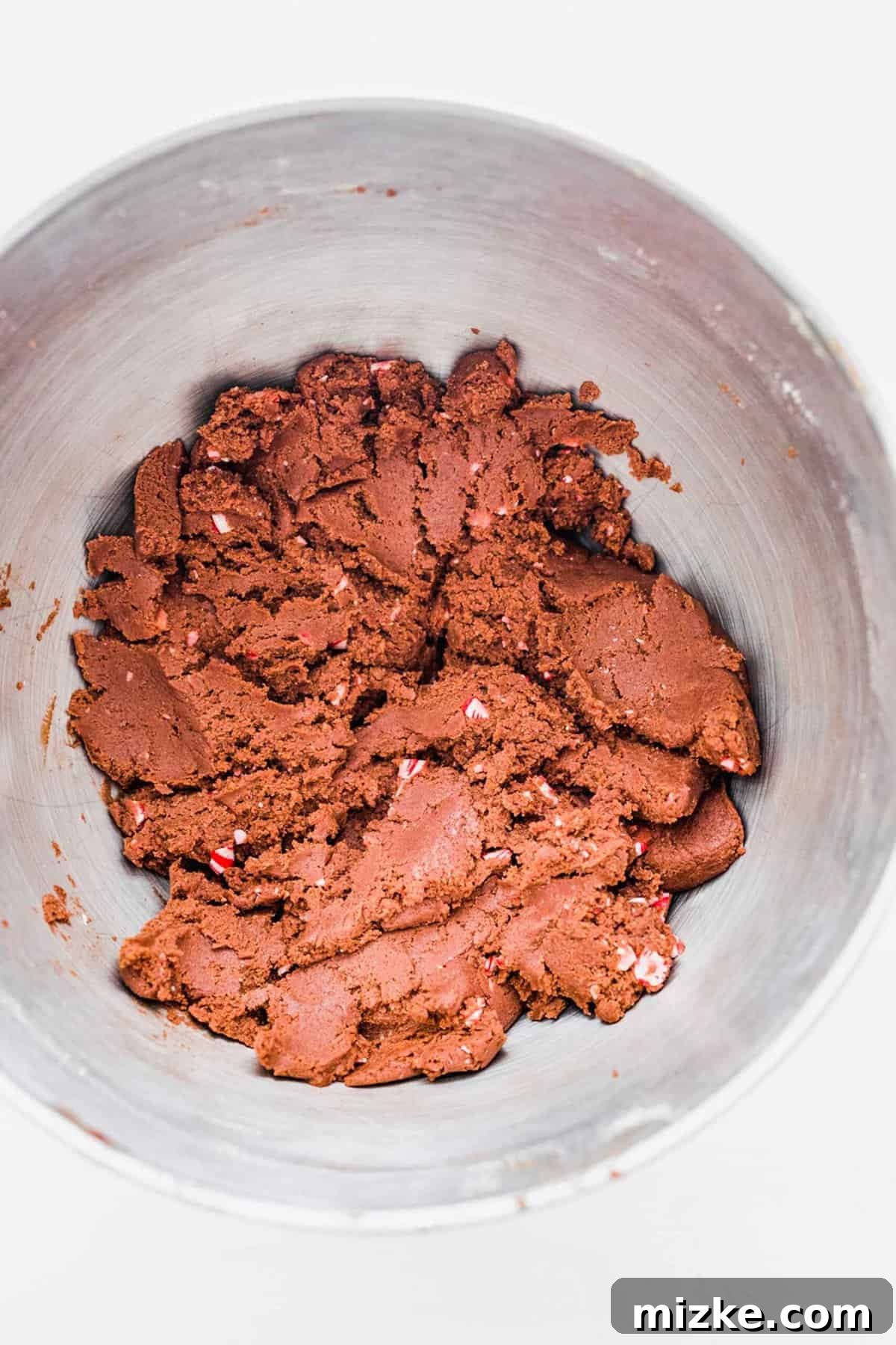 These chocolate peppermint kiss cookies are festive treats for Christmas, easy to make, and topped with a Hershey's candy cane kiss. A mixing bowl showcasing the fully incorporated chocolate peppermint kiss cookie dough, with colorful chunks of crushed candy cane visible throughout, adding texture and flavor.