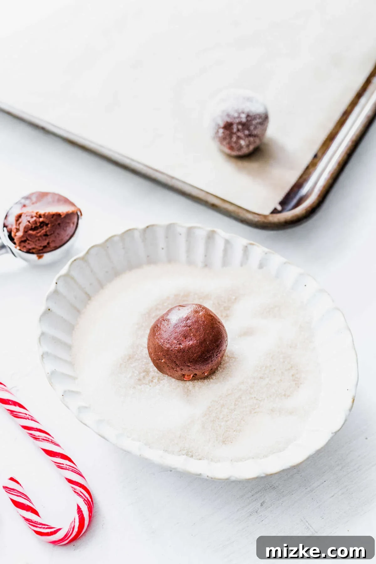 These chocolate peppermint kiss cookies are festive treats for Christmas, easy to make, and topped with a Hershey's candy cane kiss. A shallow bowl of granulated sugar with a perfectly round ball of chocolate peppermint kiss cookie dough nestled within it, ready to be coated, with a parchment-lined baking tray blurred in the background.