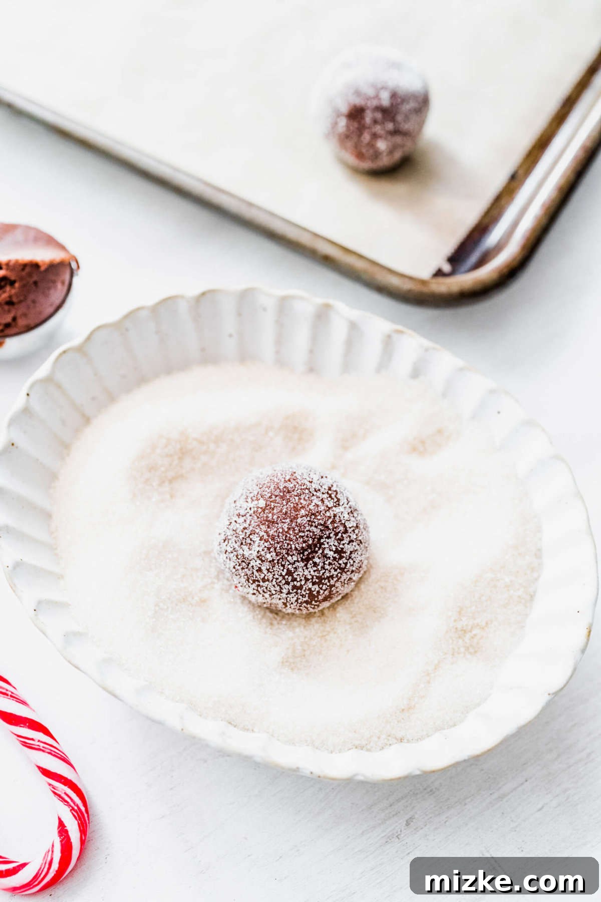 These chocolate peppermint kiss cookies are festive treats for Christmas, easy to make, and topped with a Hershey's candy cane kiss. A close-up shot of a perfectly round ball of chocolate peppermint kiss cookie dough, beautifully coated in sparkling granulated sugar, showcasing its festive readiness.
