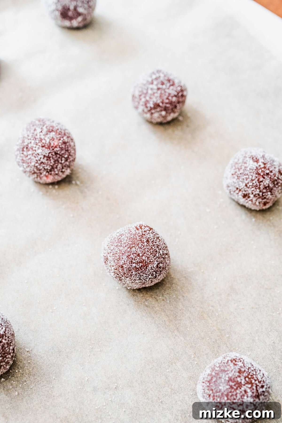 These chocolate peppermint kiss cookies are festive treats for Christmas, easy to make, and topped with a Hershey's candy cane kiss. Four perfectly portioned and sugar-coated balls of chocolate peppermint cookie dough arranged neatly on a piece of parchment paper, ready to be baked.