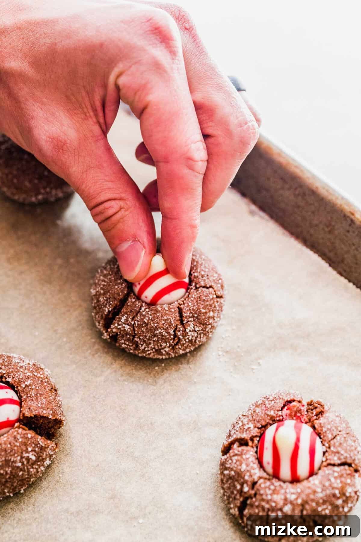 These chocolate peppermint kiss cookies are festive treats for Christmas, easy to make, and topped with a Hershey's candy cane kiss. A hand gently pushing a festive Hershey's peppermint kiss into the soft center of a freshly baked chocolate peppermint cookie, while it's still warm from the oven.