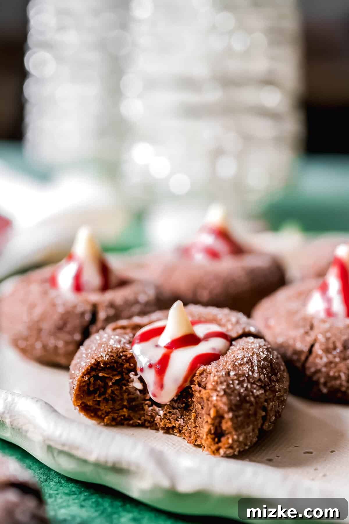 These chocolate peppermint kiss cookies are festive treats for Christmas, easy to make, and topped with a Hershey's candy cane kiss. A close-up of a chocolate peppermint kiss cookie with a perfect bite taken out, revealing its soft, rich interior, with three more untouched cookies blurred in the background.