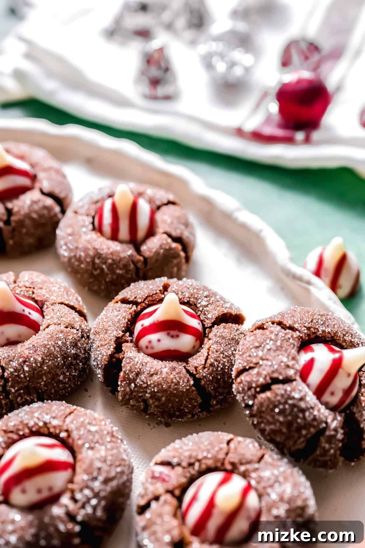 These chocolate peppermint kiss cookies are festive treats for Christmas, easy to make, and topped with a Hershey's candy cane kiss. A close-up view of several freshly baked chocolate peppermint kiss cookies, each crowned with a vibrant red and white Hershey's peppermint kiss, with more kisses blurred in the background.