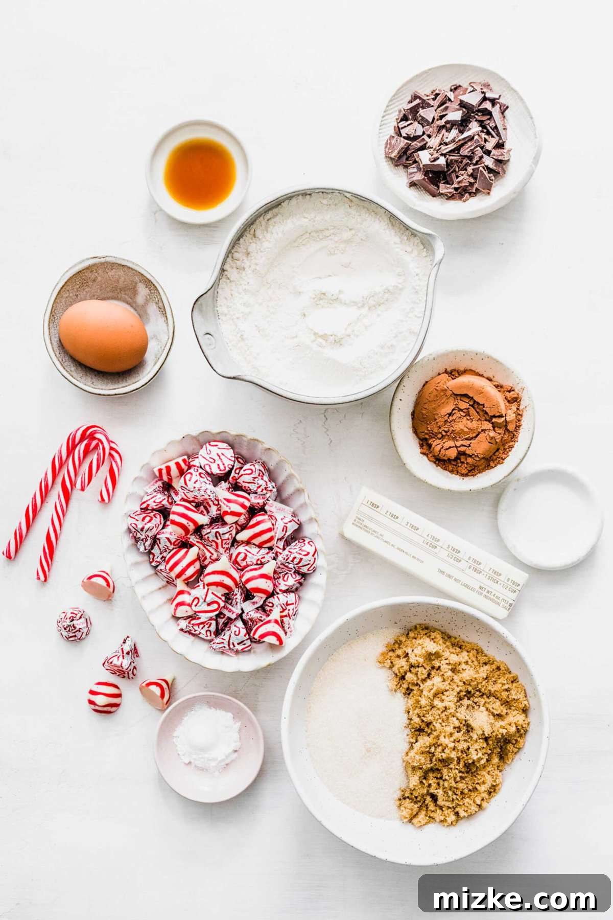 These chocolate peppermint kiss cookies are festive treats for Christmas, easy to make, and topped with a Hershey's candy cane kiss. An overhead view showcasing all the essential ingredients laid out for chocolate peppermint kiss cookies: a bowl of Hershey's peppermint kisses, a bowl of all-purpose flour, separate bowls of white and light brown sugars, a stick of unsalted butter, cocoa powder, a fresh egg, vanilla extract, some whole and crushed candy canes, peppermint extract, salt, baking powder, and semisweet chocolate pieces.