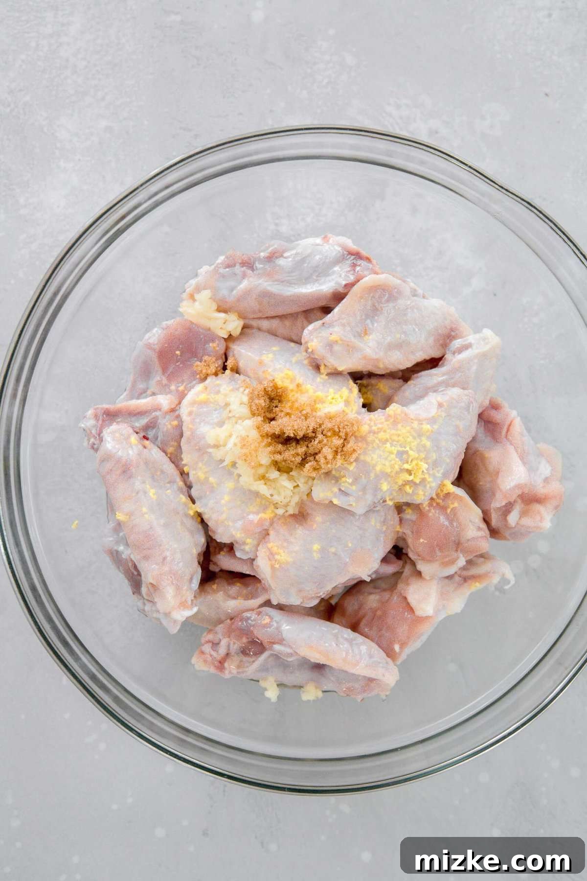 Overhead image of raw chicken wings in a large clear bowl, topped with minced garlic, grated ginger, and dark brown sugar, ready for marination.