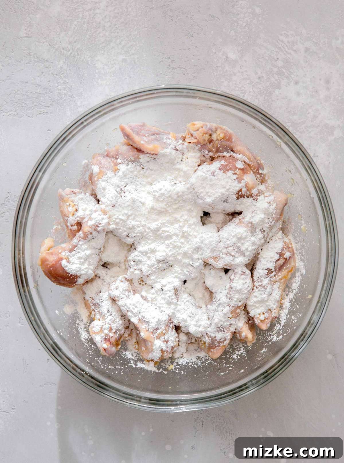 Overhead image of chicken wings in a large clear bowl with cornstarch being sprinkled generously on top, preparing for the coating step.