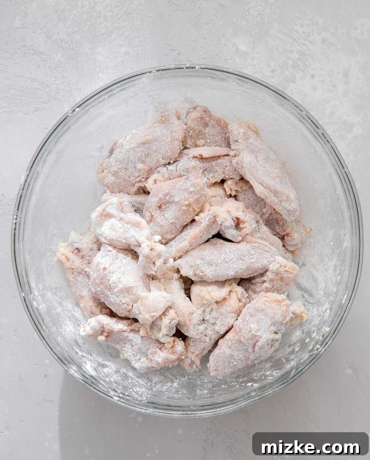 Overhead image of chicken wings thoroughly tossed in cornstarch within a large clear bowl, ensuring an even crispy coating.