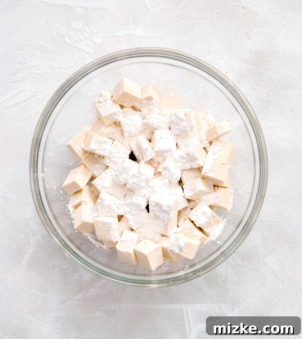 Cubed extra-firm tofu resting in a large clear glass bowl, evenly coated with a fine dusting of cornstarch.