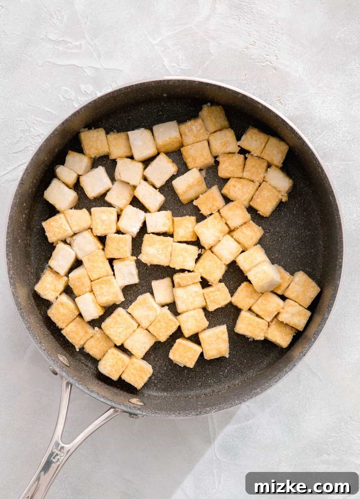 Close-up shot of cubed tofu actively pan-frying in a grey skillet, showcasing its perfectly crispy, golden-brown exterior developing as it cooks.