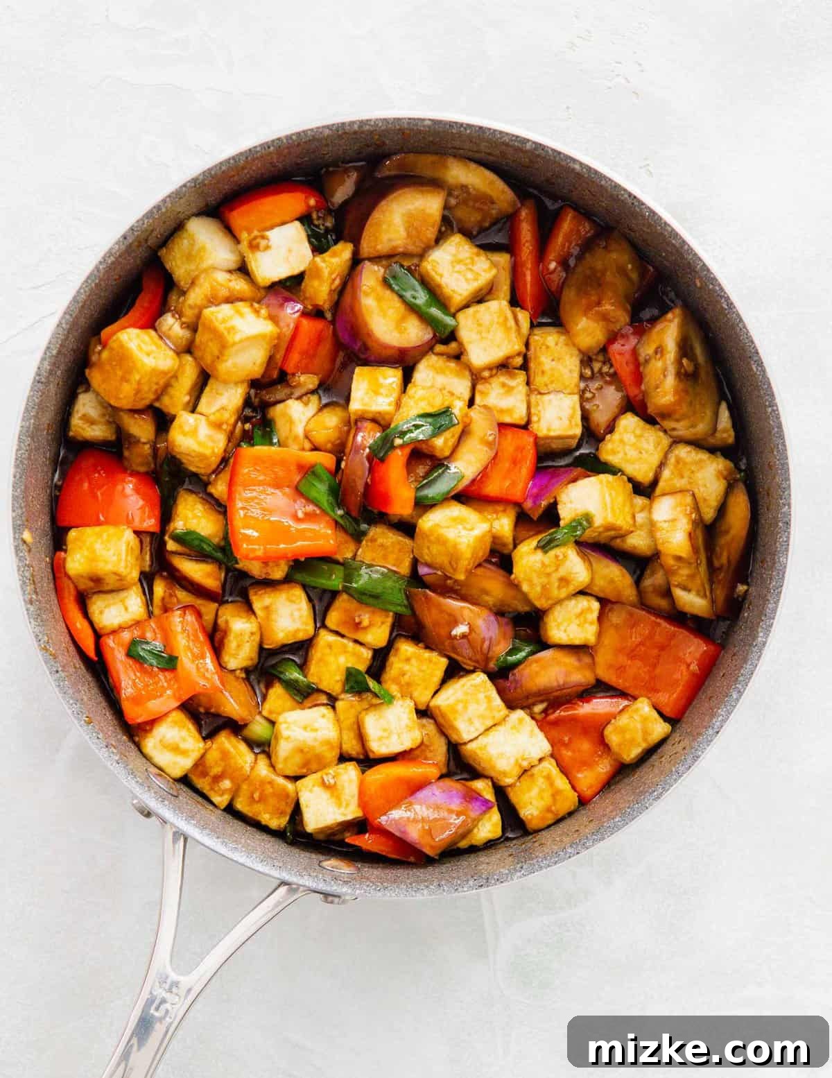 The prepared sweet and tangy sauce is poured directly into the skillet, coating the mixture of tofu, eggplant, and bell peppers, creating a glossy finish.