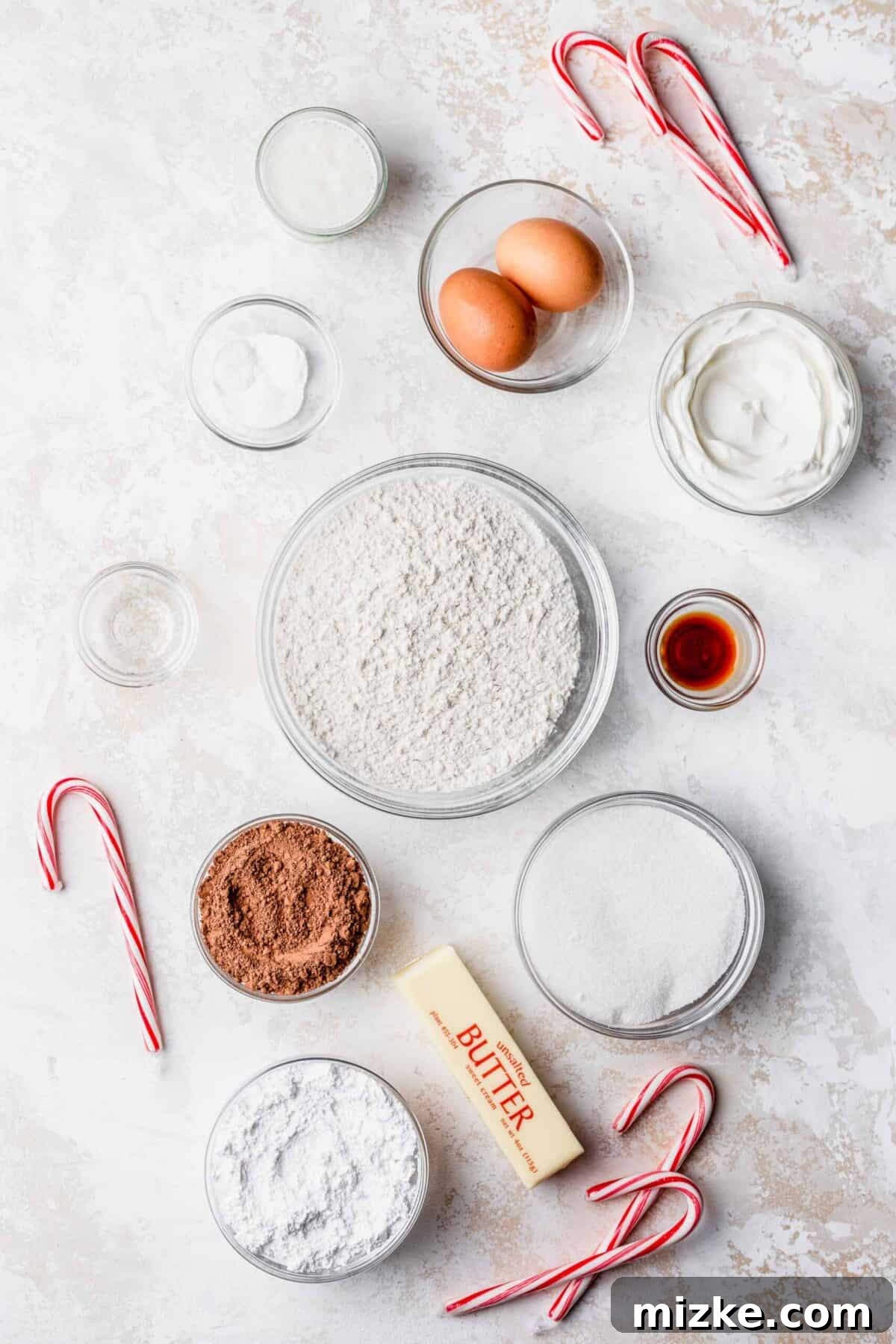 These chocolate peppermint lofthouse cookies are moist, decadent, and bursting with flavor, making them the perfect Christmas cookie. Overhead view of ingredients for chocolate peppermint lofthouse cookies: a bowl of flour, a bowl of cocoa powder, eggs, a stick of butter, candy canes, a bowl of vanilla extract, a bowl of peppermint extract, a bowl of sugar, a bowl of baking powder and baking soda, and a bowl of salt