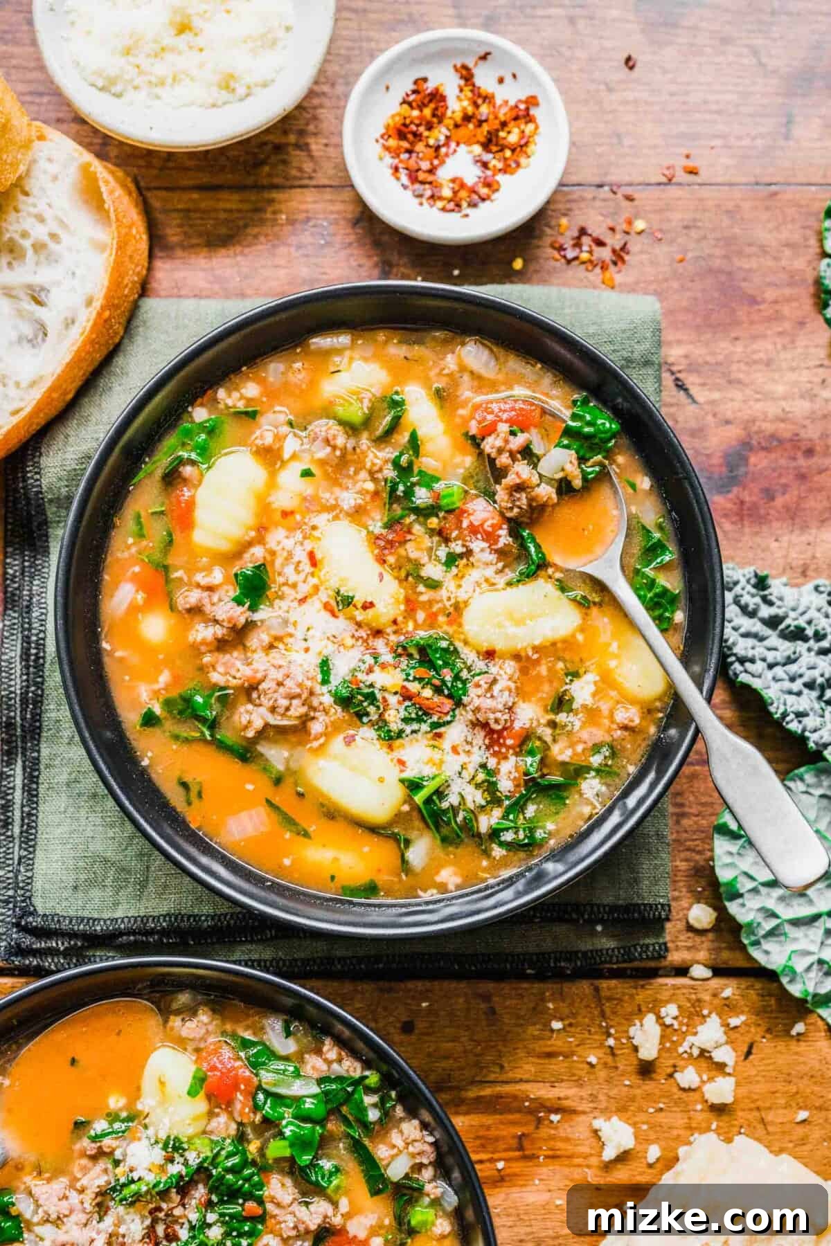Hearty Italian Sausage Gnocchi Stew 2 A steaming bowl of hearty sausage gnocchi soup with a spoon, accompanied by another bowl, fresh kale leaves, and a small dish of chili flakes.