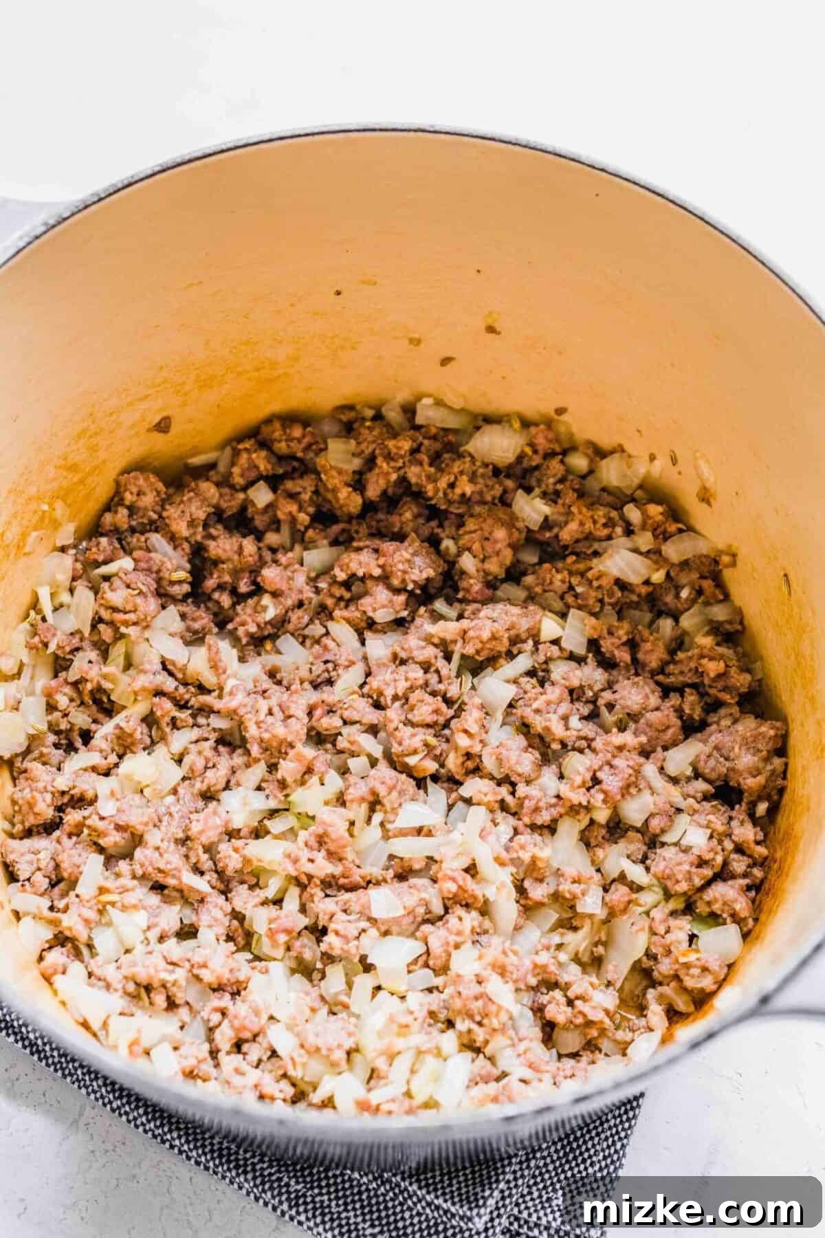 This sausage gnocchi soup recipe is comforting, spicy, and hearty, with kale, tomatoes, and lots of Italian spices. It's perfect for winter! Cooked and crumbled spicy Italian sausage mixed with softened onions in a soup pot, ready for the next ingredients.