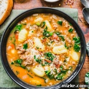 A bowl of comforting sausage gnocchi soup, garnished with parmesan, on a kitchen towel, with bread and chili flakes nearby.