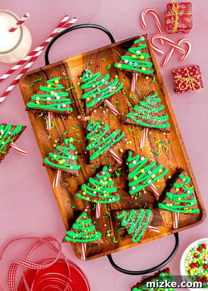 Overhead image of beautifully decorated Christmas tree brownies arranged on a rustic wooden tray, accompanied by festive faux presents, ribbons, colorful sprinkles, a glass of milk, and decorative straws, creating a cozy holiday scene.