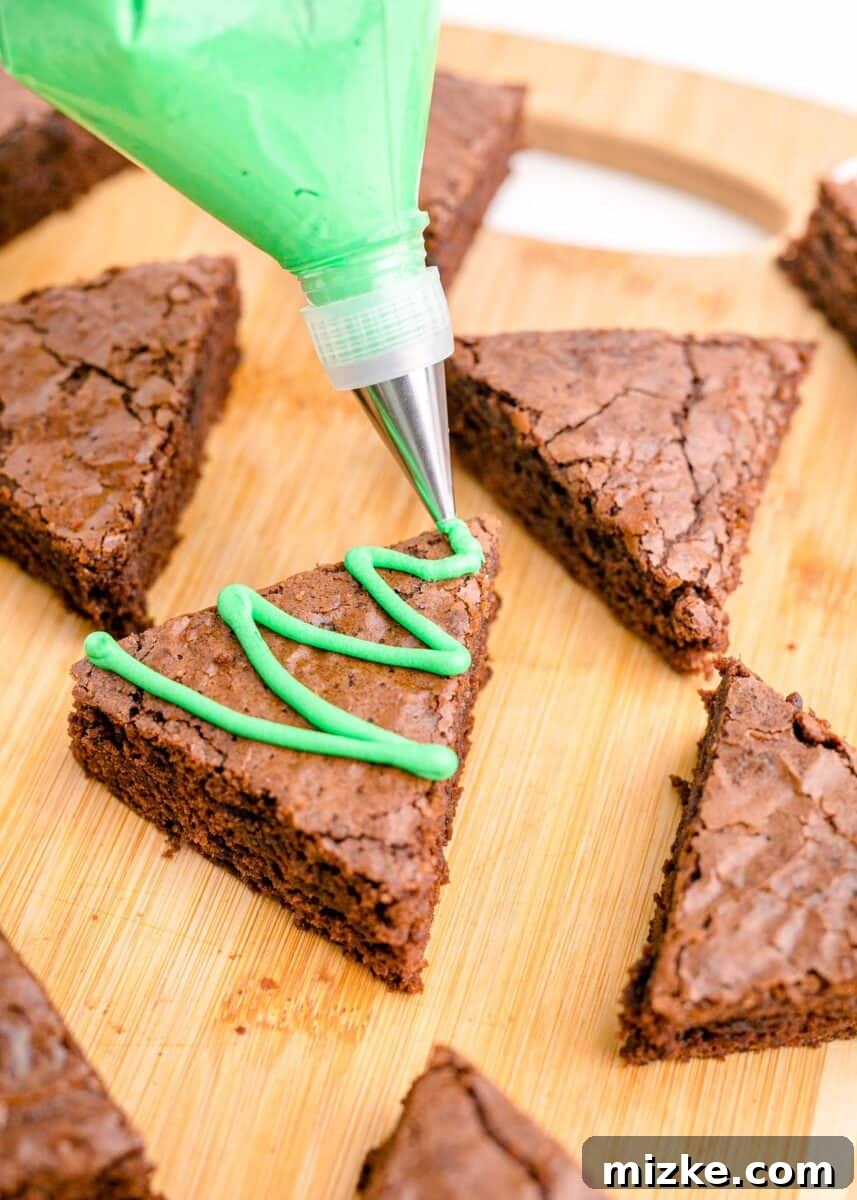 A skilled hand uses a piping bag to apply swirls of green frosting onto the tops of the brownie triangles, transforming them into festive Christmas trees.