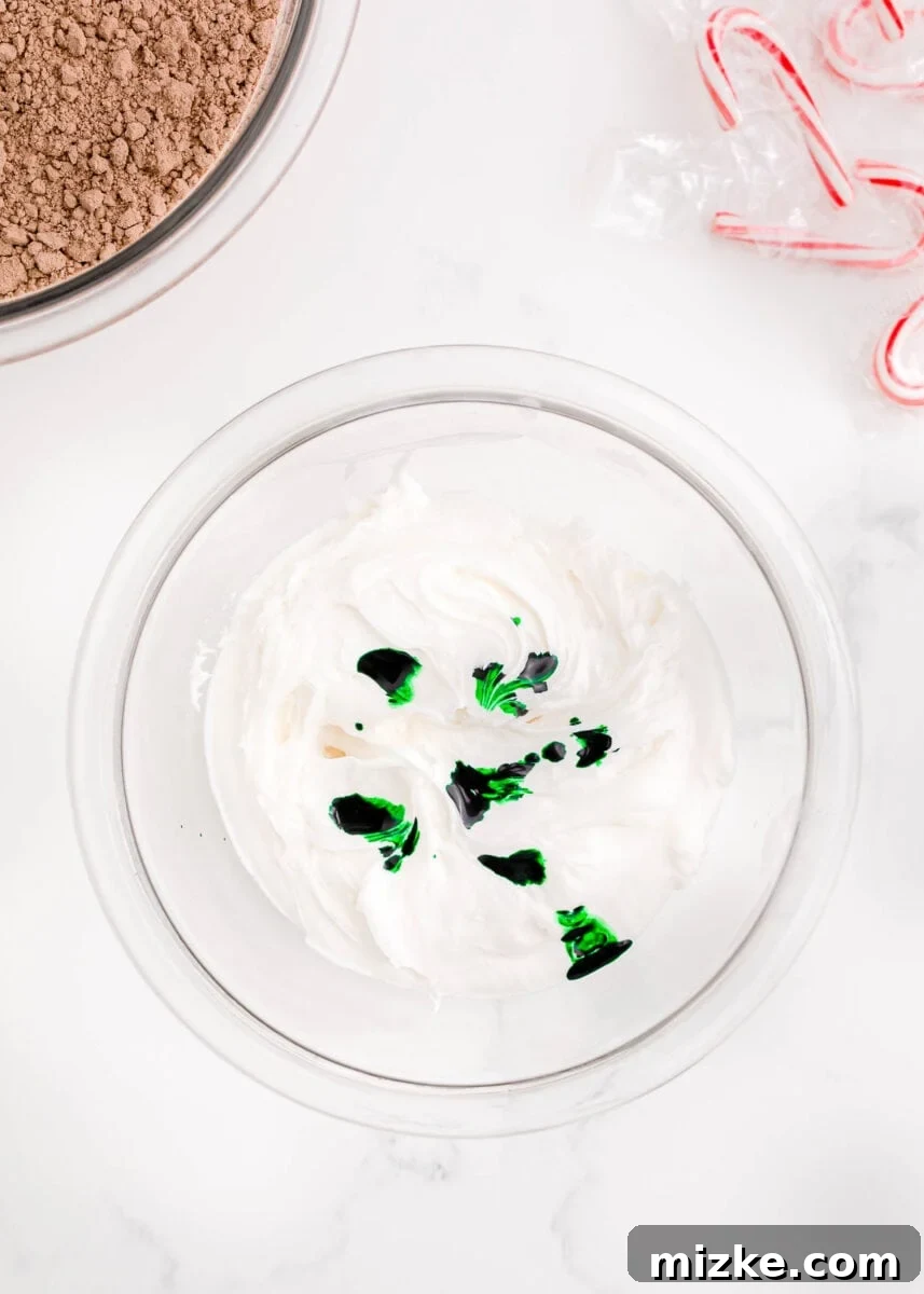 Small dots of vibrant green food coloring are carefully dropped into a bowl of creamy white frosting, alongside festive candy canes and a box of brownie mix, setting the scene for holiday decorating.
