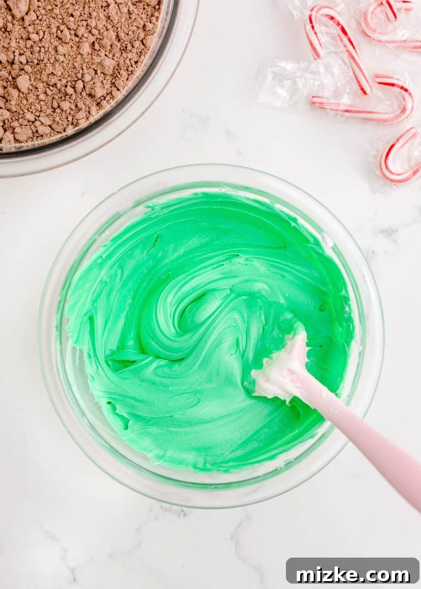 A silicone spatula skillfully blends the green food coloring into the white frosting within a clear bowl, creating a smooth, uniform green icing for decorating.