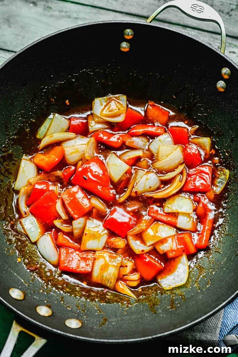 This Beijing Beef beats takeout any day. Sticky, sweet, tangy, slightly spicy sauce marries crispy bites of beef with sautéed veggies. Sautéed bell peppers and onions in a wok with Beijing beef sauce being added.