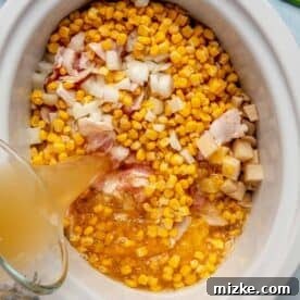 Corn kernels, diced onion, raw bacon pieces, and cubed potatoes are shown inside a slow cooker insert as chicken broth is poured over the mixture, ready for stirring.