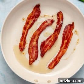 Four strips of perfectly cooked, sizzling bacon are arranged neatly in a dark cast iron skillet, showcasing their crispy texture.